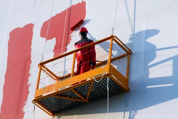 Pracownik malujący ścianę czerwoną farbą stojąc na podnośiku